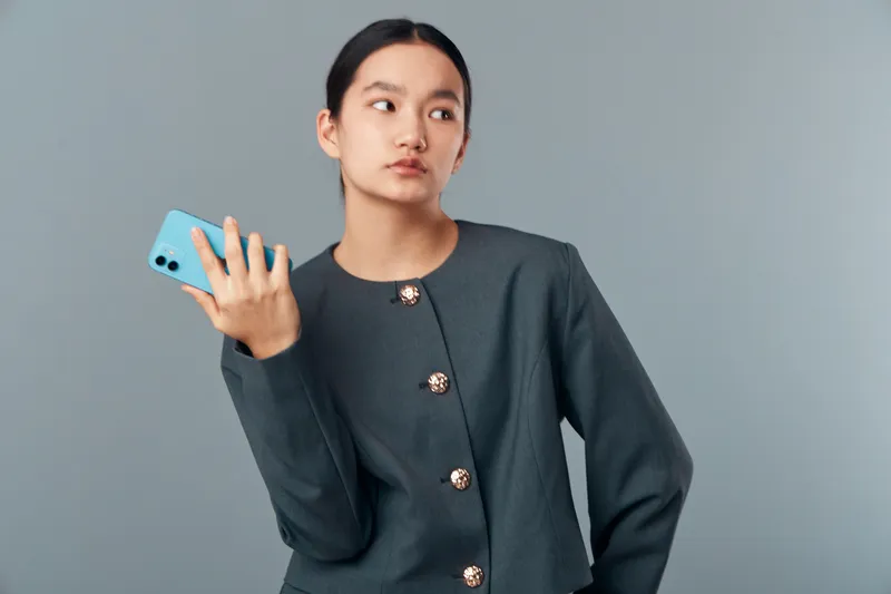 Young woman with long dark hair holding blue smartphone in hand looking away wearing dark gray buttoned jacket on plain gray background studio portrait