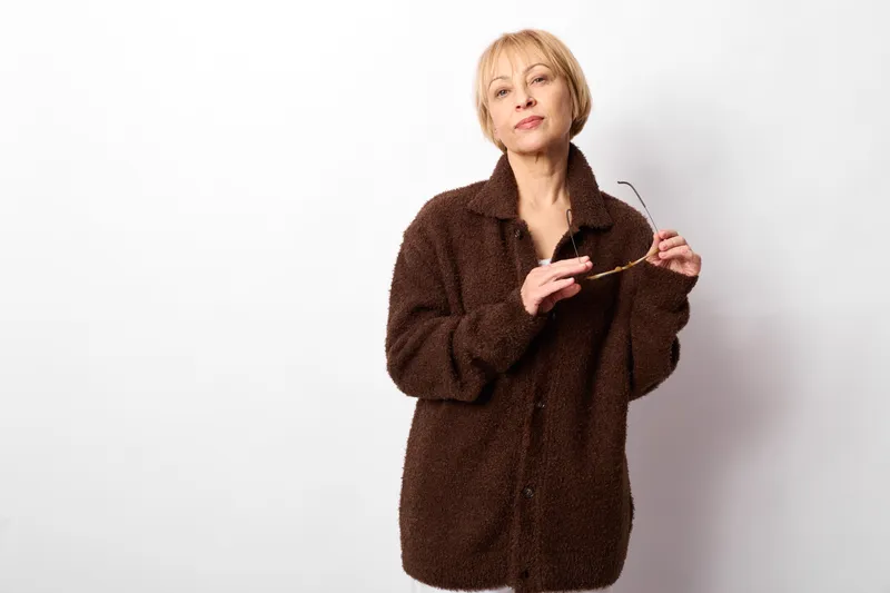 Warm elderly woman portrait, brown coat and scarf held in hands, smiling on a white studio background, cozy winter clothing, soft fur texture, comfort and self care