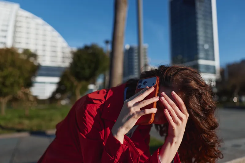 Young woman in a red jacket shields her face with a smartphone in an urban park, looking overwhelmed by a call against a city skyline.