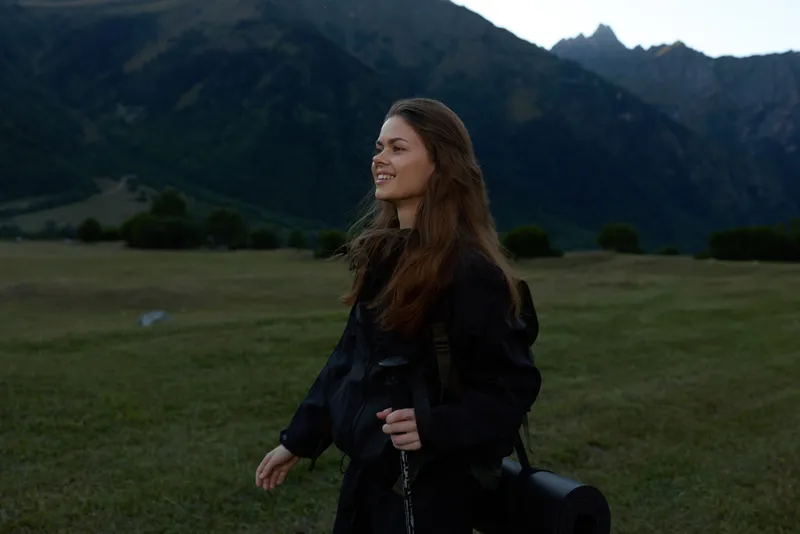 A lone woman stands in a wide grassy field beneath towering mountains, wearing a dark jacket and carrying a camera. She smiles softly, ready to capture nature's beauty.