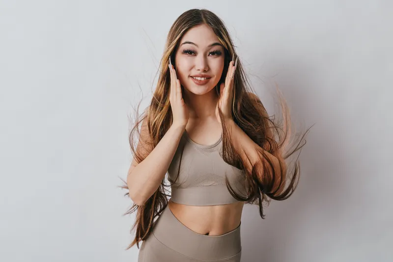 Young woman in sportswear with long hair smiling and touching face on light background. Fitness model posing with natural makeup and healthy skin for active lifestyle concept.