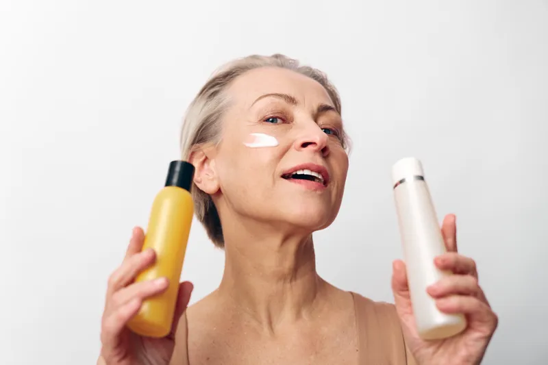 Mature woman with short blonde hair applies white cream on face holding two cosmetic bottles, smiling confidently on white background. Concept of skincare, beauty routine, and aging care.