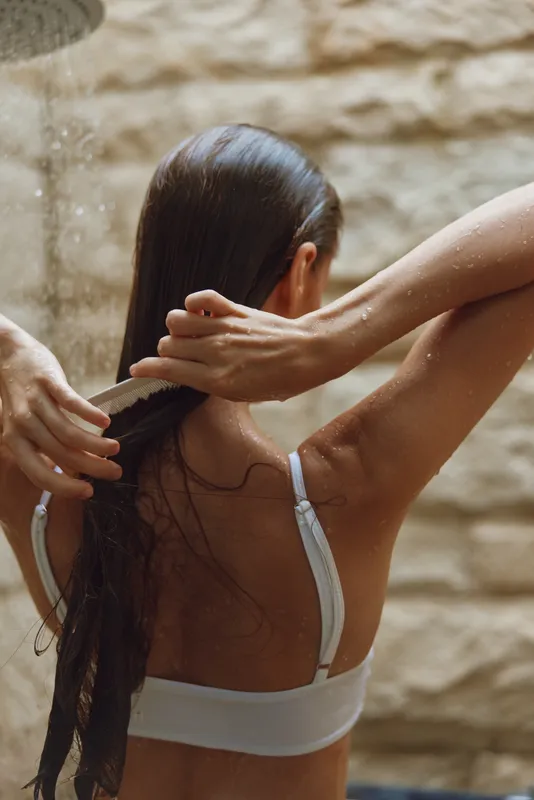 Woman washing long hair under shower with natural light, wet skin, white swimwear, and warm beige textured wall background, evoking freshness and self-care moment.