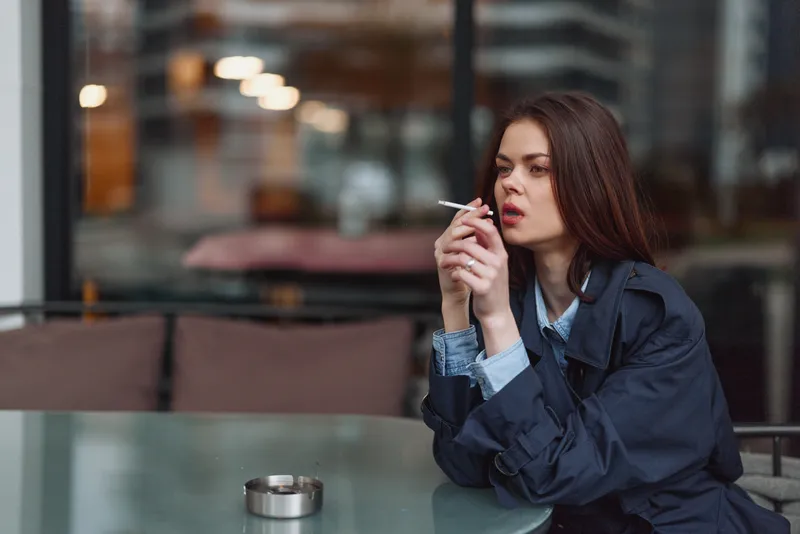 Woman sitting at an outdoor cafe smoking a cigarette, urban portrait with a contemplative mood, casual fashion, dusk lighting and mood, isolated scene.