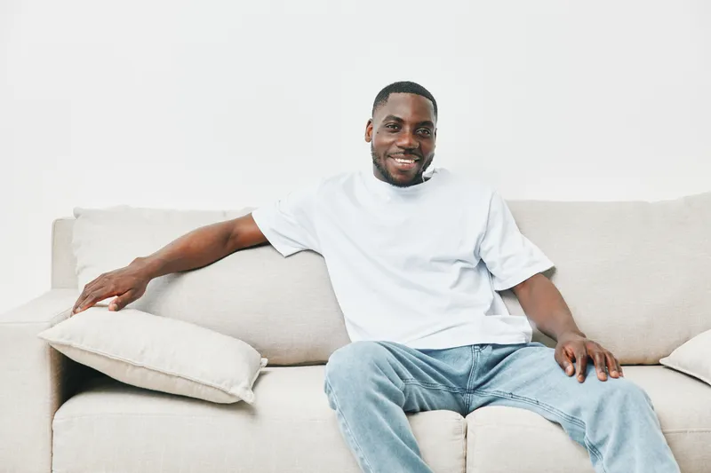 Cheerful young African American man in casual clothing relaxing on a beige sofa, conveying a sense of comfort and happiness