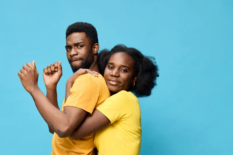 Young African American couple in yellow shirts embracing against a vibrant blue background, expressing joy and connection with playful emotions