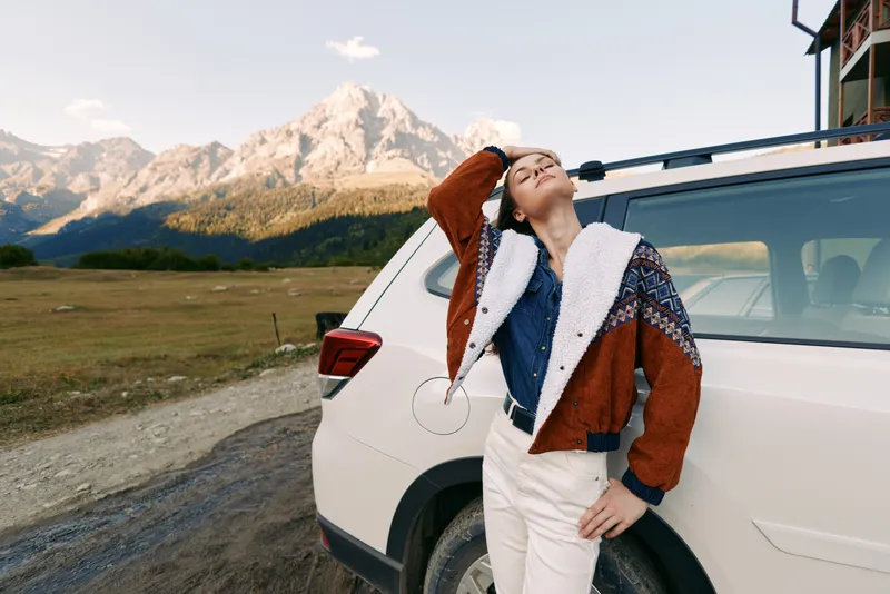 Woman leaning on car by mountains during travel, enjoying nature and open road outdoors, relaxed pose in cozy sweater, lifestyle vacation moment with scenic alpine backdrop.