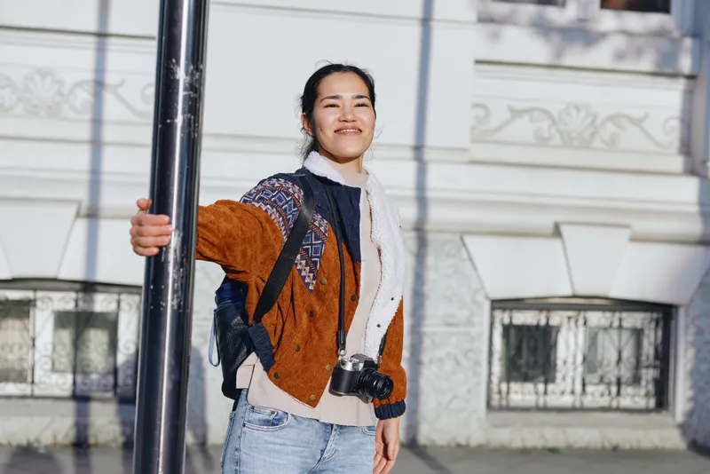 Stylish woman outdoors in a fashionable outfit with scarf, denim jeans, and a warm jacket beside a street post, smiling in an urban moment