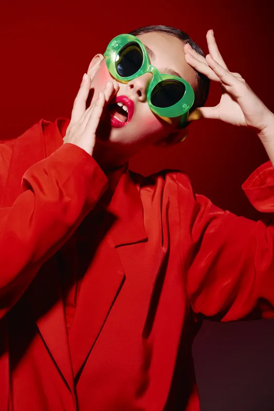 Fashion model in oversized green sunglasses and a bold red coat poses with an expressive face on a vivid red background. Creative beauty and modern style concept with striking colors.