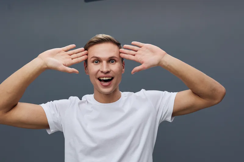 Excited happy young caucasian man wear white t-shirt gesture open hands near head look camera isolated on grey background facial expression concept digital detox mental health