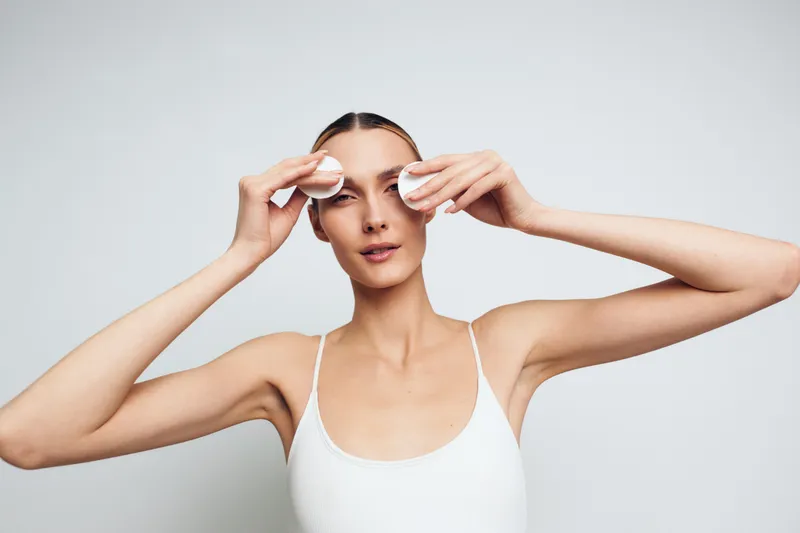 Young woman in white tank top removing makeup with cotton pads on light background. Concept of skincare, beauty routine, and facial cleansing with natural light.