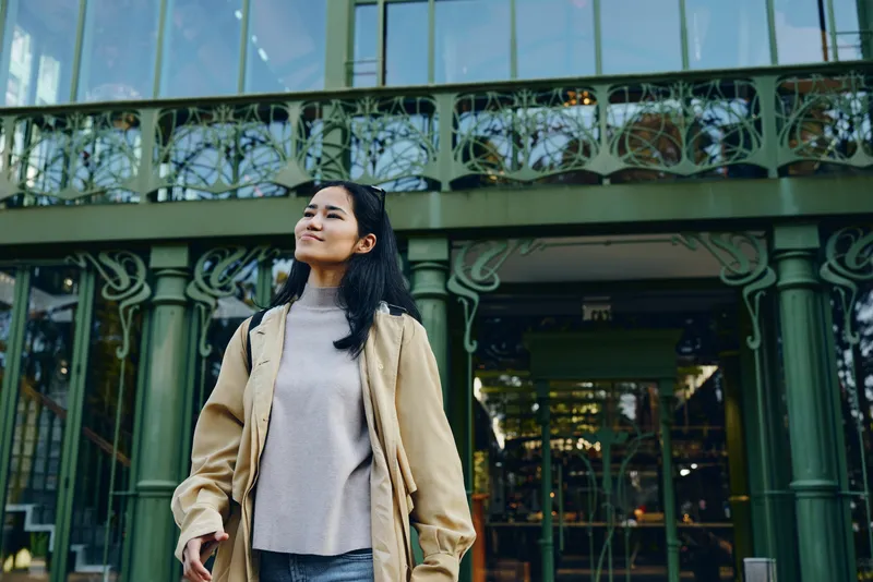 Woman outdoor fashion coat urban confident standing in front of an ornate glass and iron building, stylish candid scene showcasing contemporary lifestyle and chic streetwear