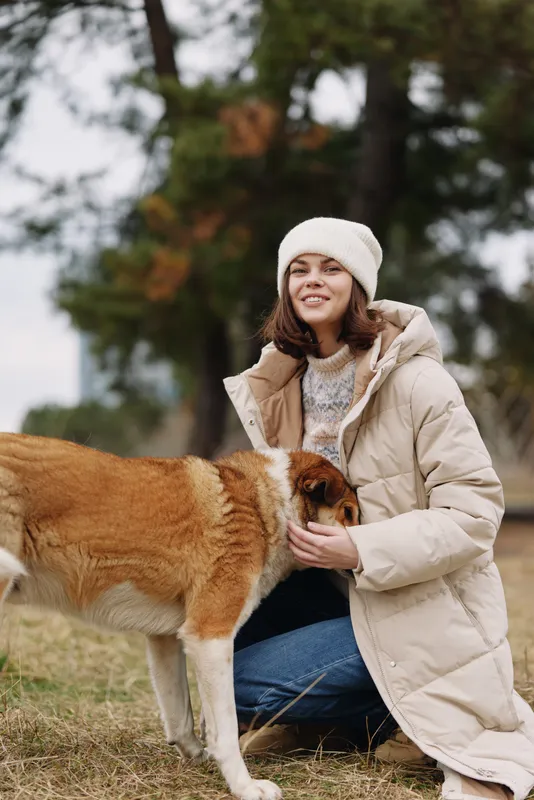 Dog and woman in an outdoor setting, wearing a warm coat and beanie, sharing a gentle moment of interaction and care in a tranquil countryside