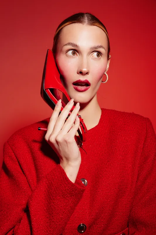 Young woman in red coat holds red high heel shoe near face with surprised expression on red background. Fashion concept with bold colors and stylish accessories.