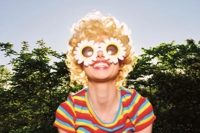 Playful outdoor portrait of a smiling woman in retro film colors, wearing oversized flower sunglasses, striped shirt, and natural greenery behind, casual lifestyle moment on bright day.