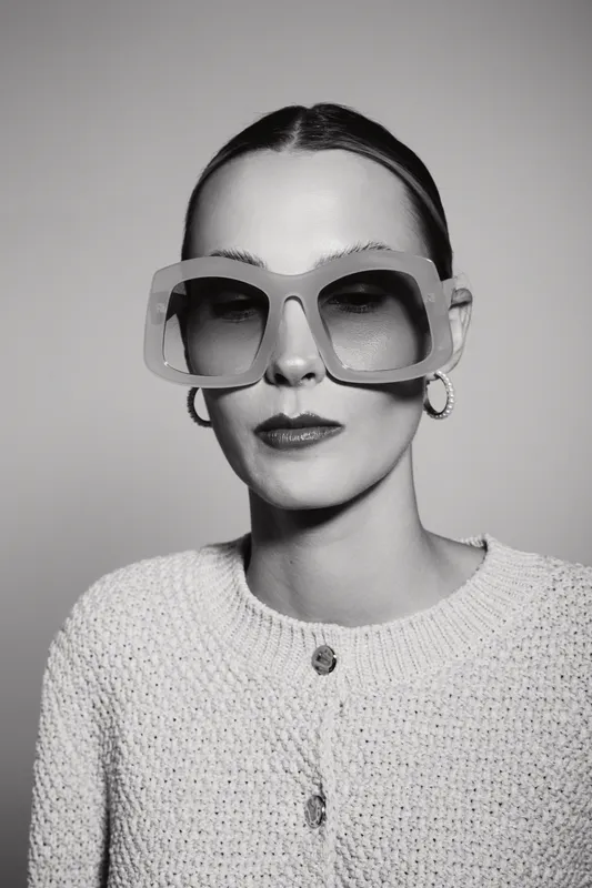 Stylish Caucasian woman with pale skin and slicked-back hair wears oversized sunglasses and knitted sweater in a studio. Concept of fashion, elegance, and modern style.
