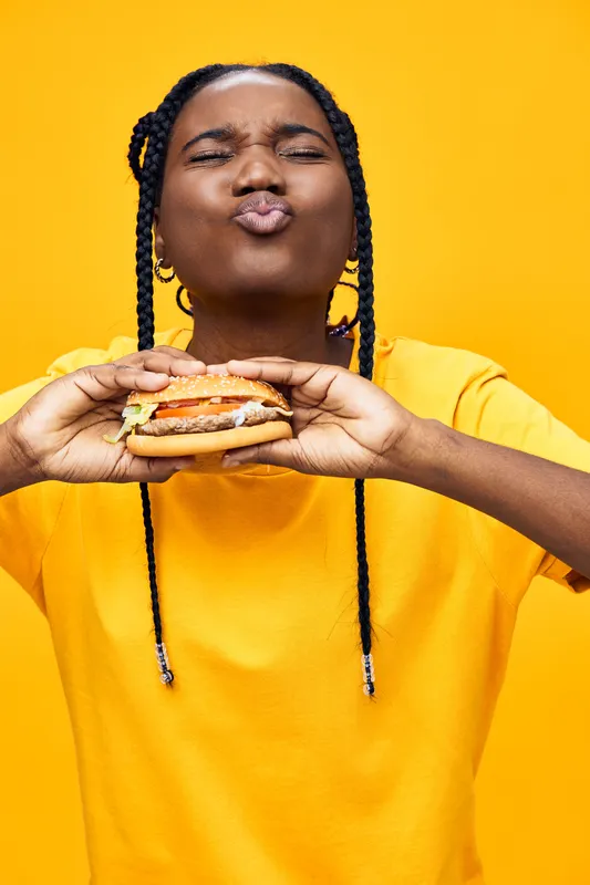 A joyful young woman with braids savoring a hamburger against a vibrant yellow background, expressing happiness and delight in fast food indulgence