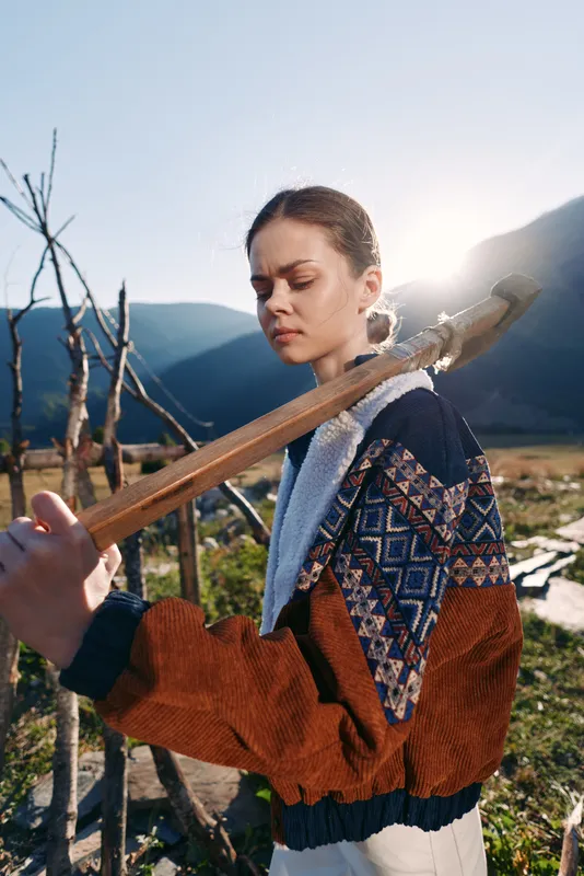 Woman portrait outdoors in mountains at sunrise carrying a wooden farming tool on her shoulder, rural countryside scene with traditional jacket, contemplative expression and sunlight.