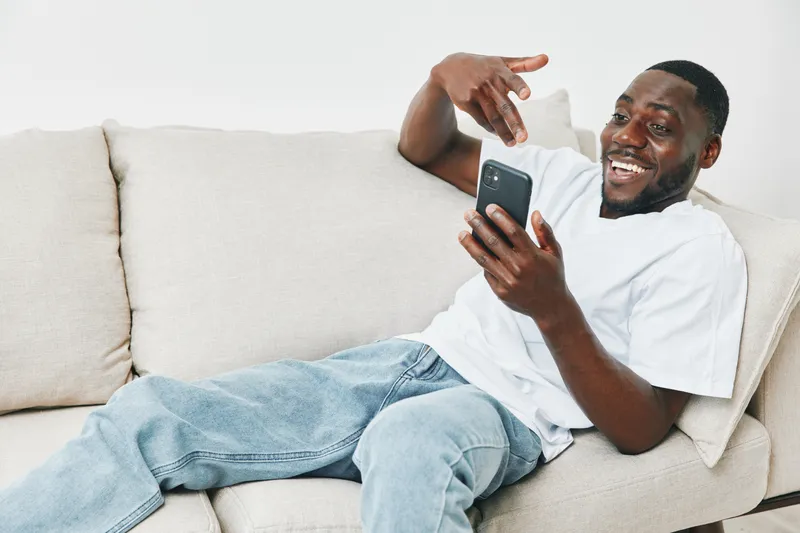 Happy young African American man using smartphone while relaxing on couch at home, enjoying social media moments and laughter, cozy atmosphere, casual style