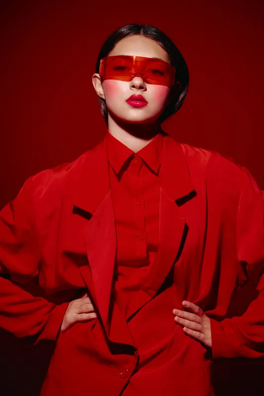 Young woman in red oversized jacket and matching red sunglasses poses confidently against red background. Concept of beauty, fashion, and creative style with vibrant color and attitude.