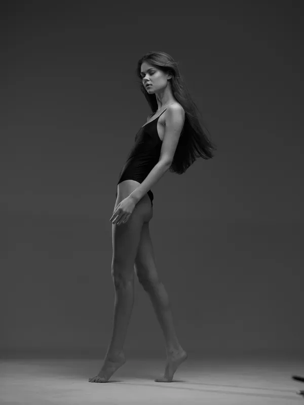 Slim woman walking in black leotard with flowing hair on monochrome background, graceful pose, elegant movement.