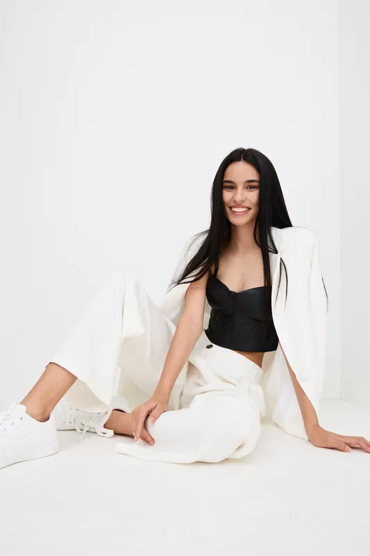 smiling woman wearing stylish white suit and black top sitting on floor with long dark hair and natural makeup in minimalistic white room interior