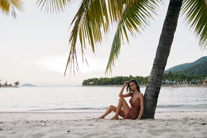 Young woman relaxing under palm tree on tropical beach during sunset with calm sea and distant mountains in background.