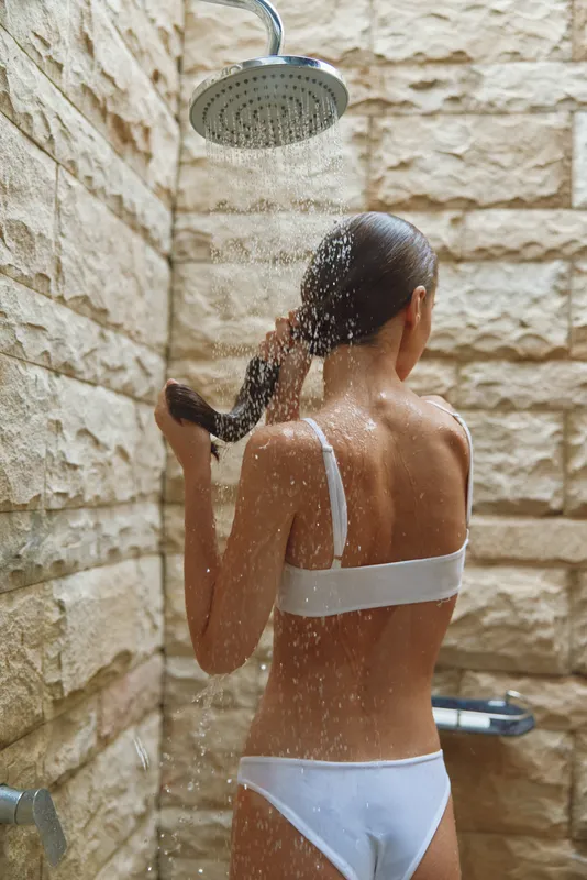 Woman showering under rain shower head in stone-walled bathroom, wearing white underwear and holding hair, refreshing and cleansing moment.