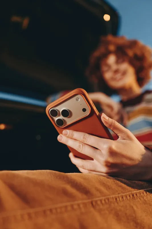 Woman lifestyle, hands holding smartphone on bed, red case, warm indoor light, blurred woman in background, communication, modern technology, cozy evening relaxing moment at home, comfort