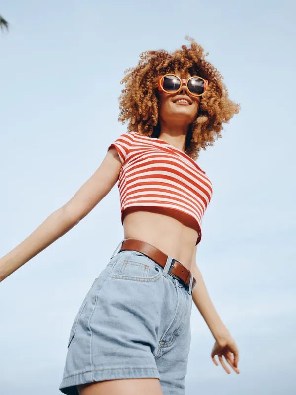 Young woman wearing a red and white striped crop top, denim high waist shorts, brown belt, and round sunglasses, posing joyfully against a bright blue sky.