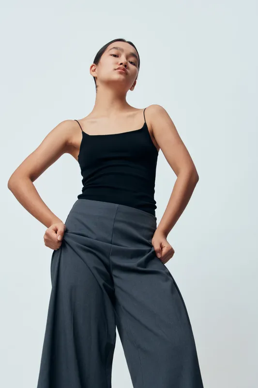 Woman standing in black tank top and wide gray pants posing confidently with hands in pockets against plain background. Young female model with serious expression and casual style.
