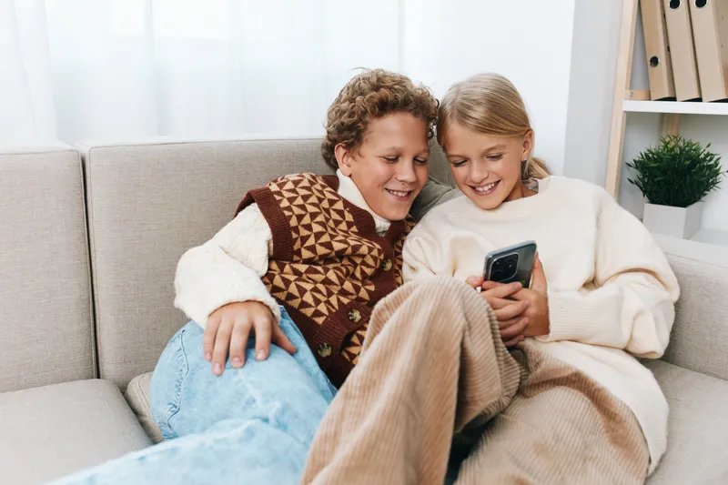 Two smiling children sit close together on a cozy sofa, sharing a mobile device and enjoying a warm, playful moment at home, surrounded by soft daylight and relaxed atmosphere.