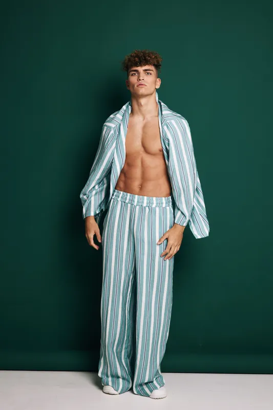 Portrait of young man with curly hair wearing unbuttoned striped pajama set showing toned abs and confident expression on green background in white shoes standing upright.
