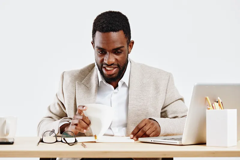 Professional young African American man working at a modern desk with laptop, coffee mug, and glasses, dressed in beige blazer and white shirt, smiling and focused on his tasks, indoor office environm