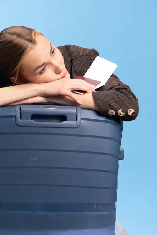 Young woman leans on large blue suitcase holding white boarding pass, looking thoughtful. Concept for travel, waiting, and planning on blue background with casual clothing and relaxed mood.