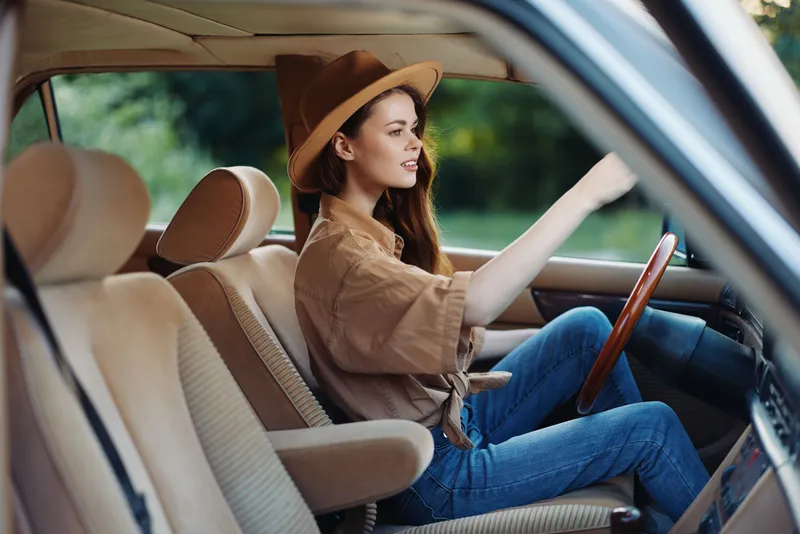 Stylish young woman in a car, wearing a brown hat and casual outfit, exuding confidence and joy against a blurred green background.