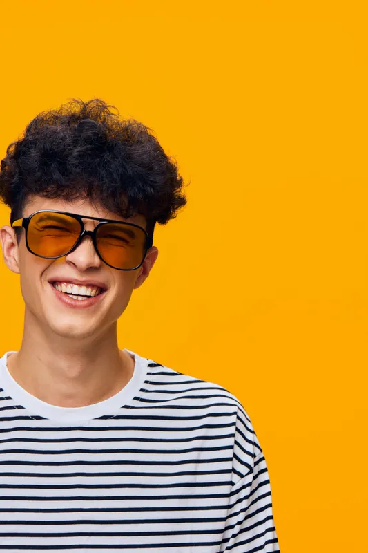 smiling man with sunglasses wearing striped shirt on vibrant yellow background expressing happiness and youthfulness with curly hair and casual style