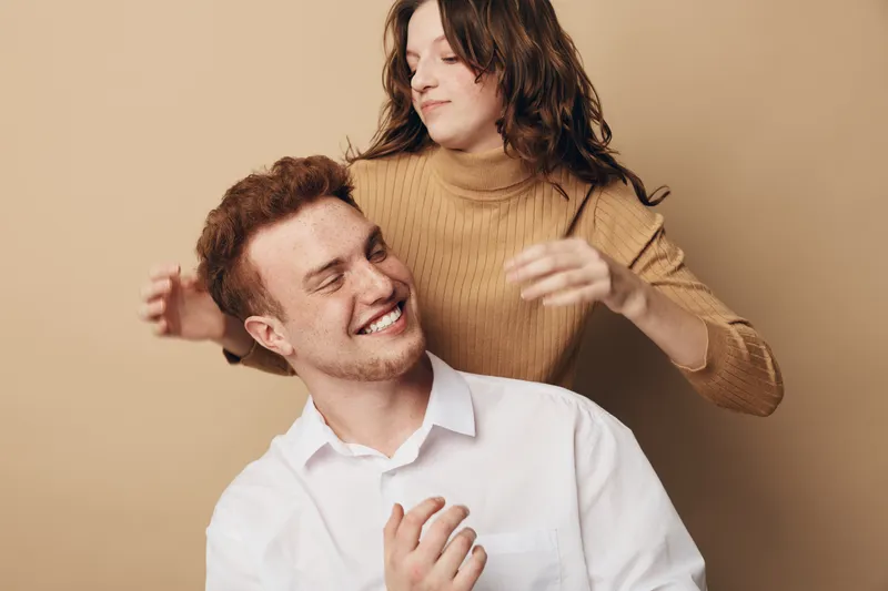 Happy young couple in casual clothes enjoys playful moment together on beige background. Smiling man and woman express joy and affection in warm studio setting.