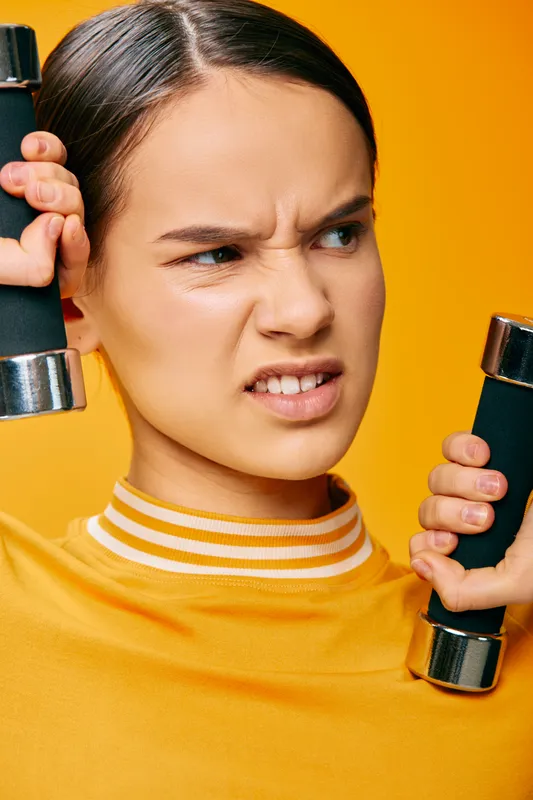 Female fitness athlete in a yellow gym top holding dumbbells, focused expression, mid workout, strong posture, dynamic training scene in a modern studio.