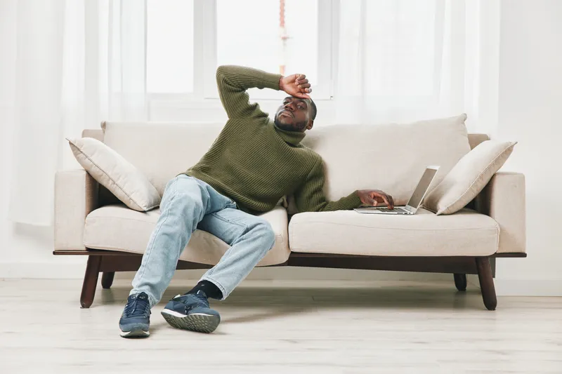 Tired young African American man in casual clothes relaxing on a couch with a laptop, expressing fatigue in a bright living room setting