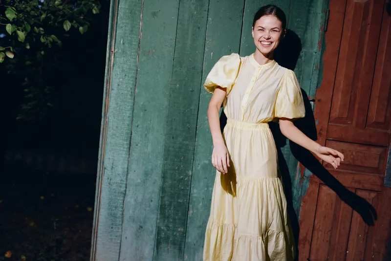 woman smile in yellow dress outdoors in sunlight against rustic wooden wall portrait with authenticity inclusive and happy