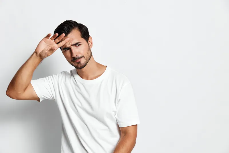 Stylish man in a white T-shirt posing with a hand salute gesture, expressing confidence and charm against a minimalistic light gray background. People lifestyle concept