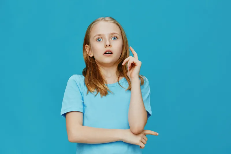 Surprised girl in a blue t shirt with light brown hair on a bright blue background showing a curious expression, ideal for concepts of childhood or wonder
