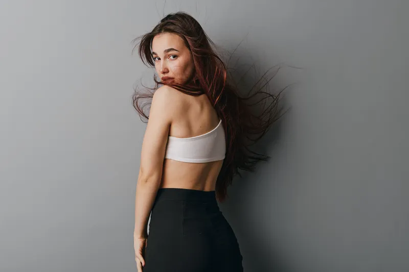 Young woman with long hair in white top and black pants posing against gray wall. Female model looking over shoulder with natural makeup and casual style in studio lighting.