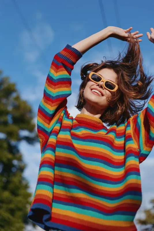 Woman in rainbow sweater and sunglasses smiling outdoors, portrait of happy young woman raising arms, colorful knitwear, joyful fashion and sunny casual style.