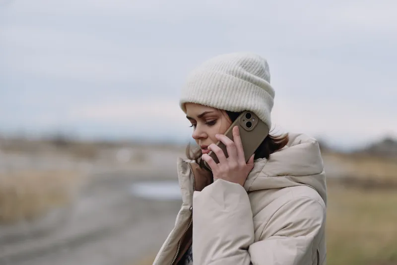 Woman in a warm beige coat and white beanie speaks on her cellphone outdoors in a brisk landscape, conveying calm focus, clear communication and solitude during a cold, quiet day.