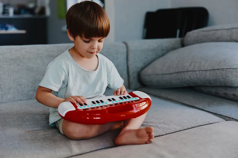 Young boy playing a red keyboard, sitting on a cozy sofa in a stylish living room, showcasing creativity and joy with a focus on music and fun.