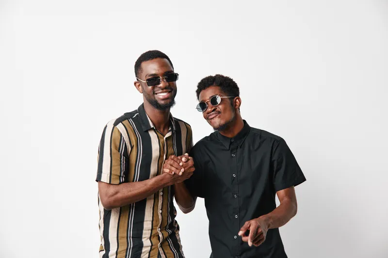 Two African American men with dark skin wearing sunglasses smiling and shaking hands in casual shirts on white background for friendship and connection.
