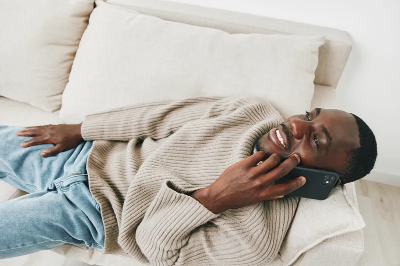 Smiling African man talking on the phone while relaxing on a couch, wearing a cozy sweater and jeans, showcasing a warm and inviting atmosphere
