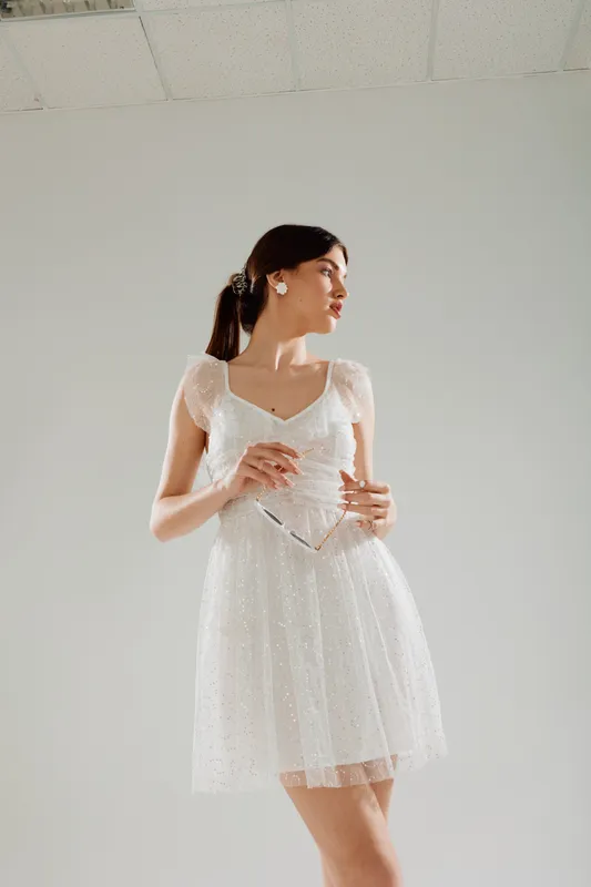 Young woman in a white dress holding sunglasses looks sideways in a bright studio. Concept of woman beauty fashion and elegance with soft natural lighting.